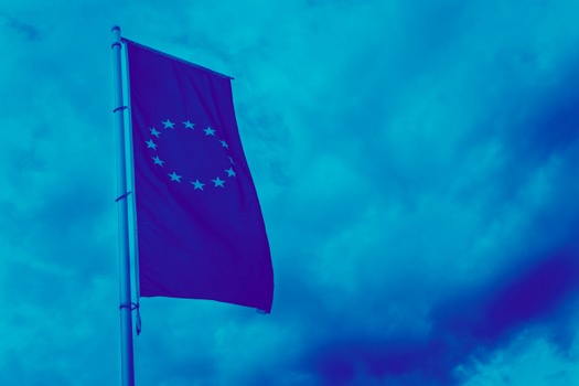 Sinisävyinen kuva, jossa EU-lippu salossa, taustalla pilvinen taivas 
