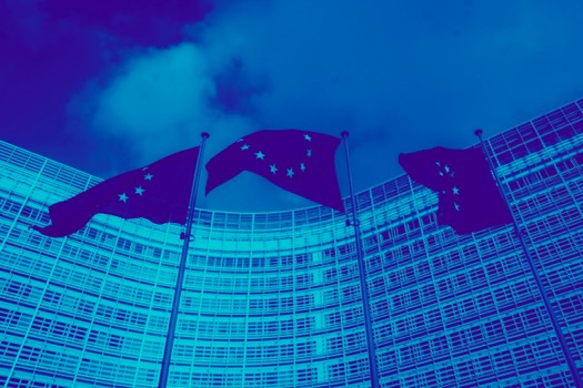 Sinisävyinen kuva, jossa kolme EU-lippua salossa, taustalla hallintorakennus ja pilvinen taivas 