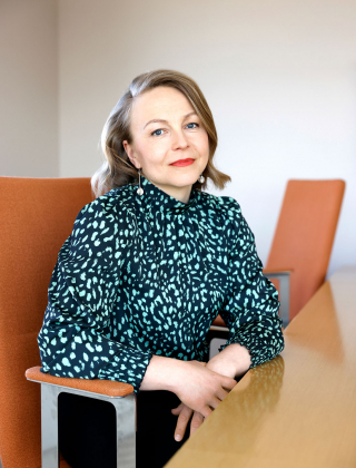Johtava asiantuntija Annukka Saari istuu oranssilla tuolilla pöydän ääressä ja katsoo kameraan.