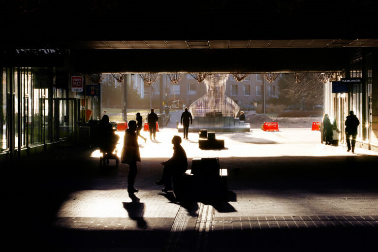 Ihmisiä kävelemässä ja istumassa varjoisalla käytävällä, taustalla auringonvalossa näkyvä veistos.