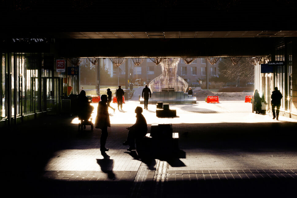 Ihmisiä kävelemässä ja istumassa varjoisalla käytävällä, taustalla auringonvalossa näkyvä veistos.