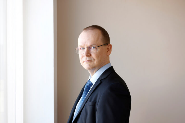 Silmälasipäinen, vakava Jarkko Ruohoniemi seisoo sivuttain puku päällä ikkunan äärellä ja katsoo kameraan.