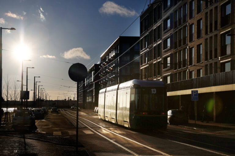 Raitiovaunu ajaa kirkkaassa auringonvalossa modernien kerrostalojen edustalla.