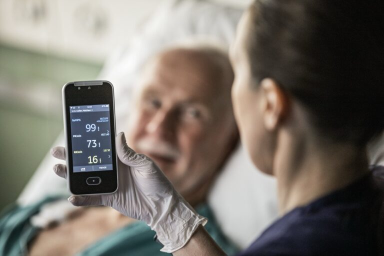 GE Healthcaren potilasmonitori hoitajan kädessä, taustalla potilas.