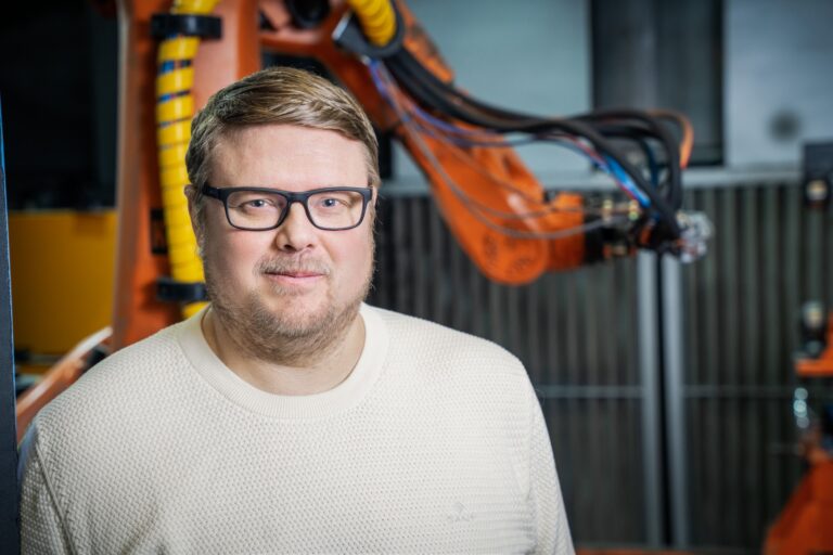 Kuopion konepajan toimitusjohtaja Jukka Finne lähikuvassa, taustalla oranssi mekaaninen työlaite.