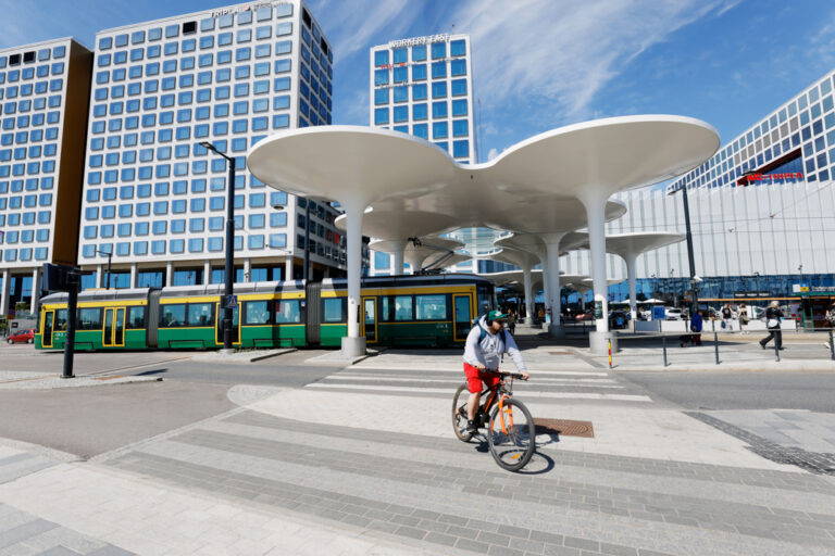 Mies pyöräilee Triplan edustalla aurinkoisessa keslissä, taustalla on raitiovaunu.