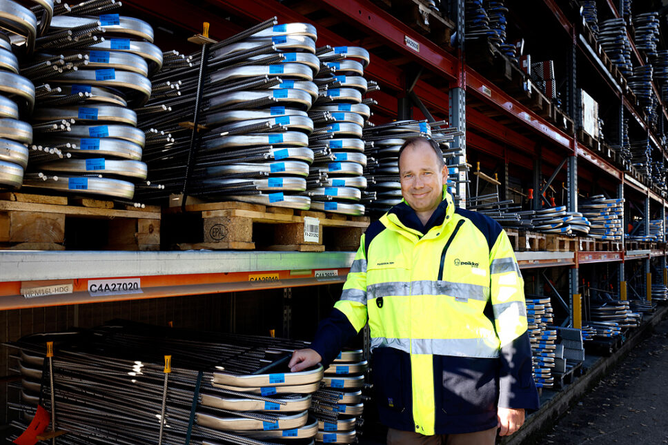 Peikko Groupin toimitusjohtaja Topi Paananen eisoo keltaisessa työtakissa tehtaalla, taustakka hyllyillä teräslieriöitä.