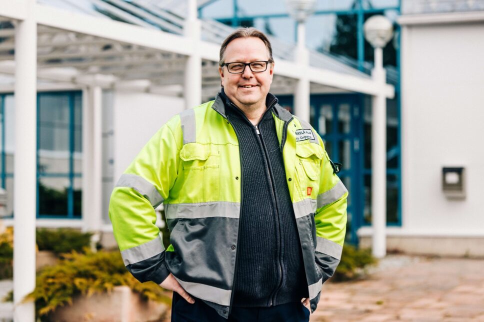 Mies seisoo vaalean rakennuksen edessä keltaharmaa huomiotakki päällä ja hymyilee.