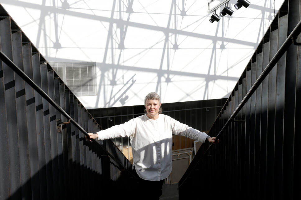 Lyhythiuksinen nainen valkoisessa paidassa seisoo kapeassa, valoisassa portaikossa pitäen käsiään kaiteilla.