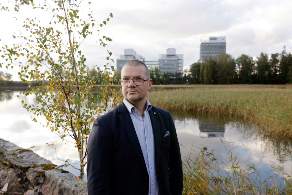 Mies seisoo hoikan koivun edessä, taustalla kaislikkoinen lahti, jonka takana rakennuksia.