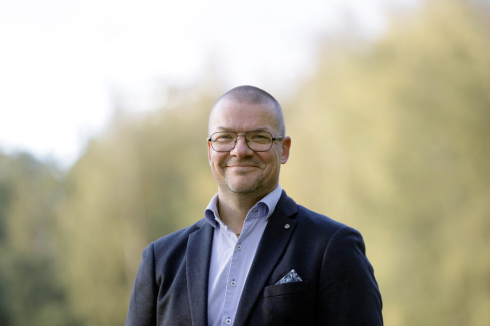 Silmälasipäinen mies hymyilee kameralle, taustalla näkyy epätarkasti luontoa.