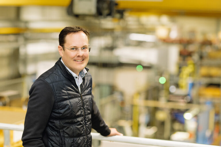 Silmälasipäinen mies mustassa takissa katsoo kameraan ja hymyilee, taustalla epätarkkanan konesali